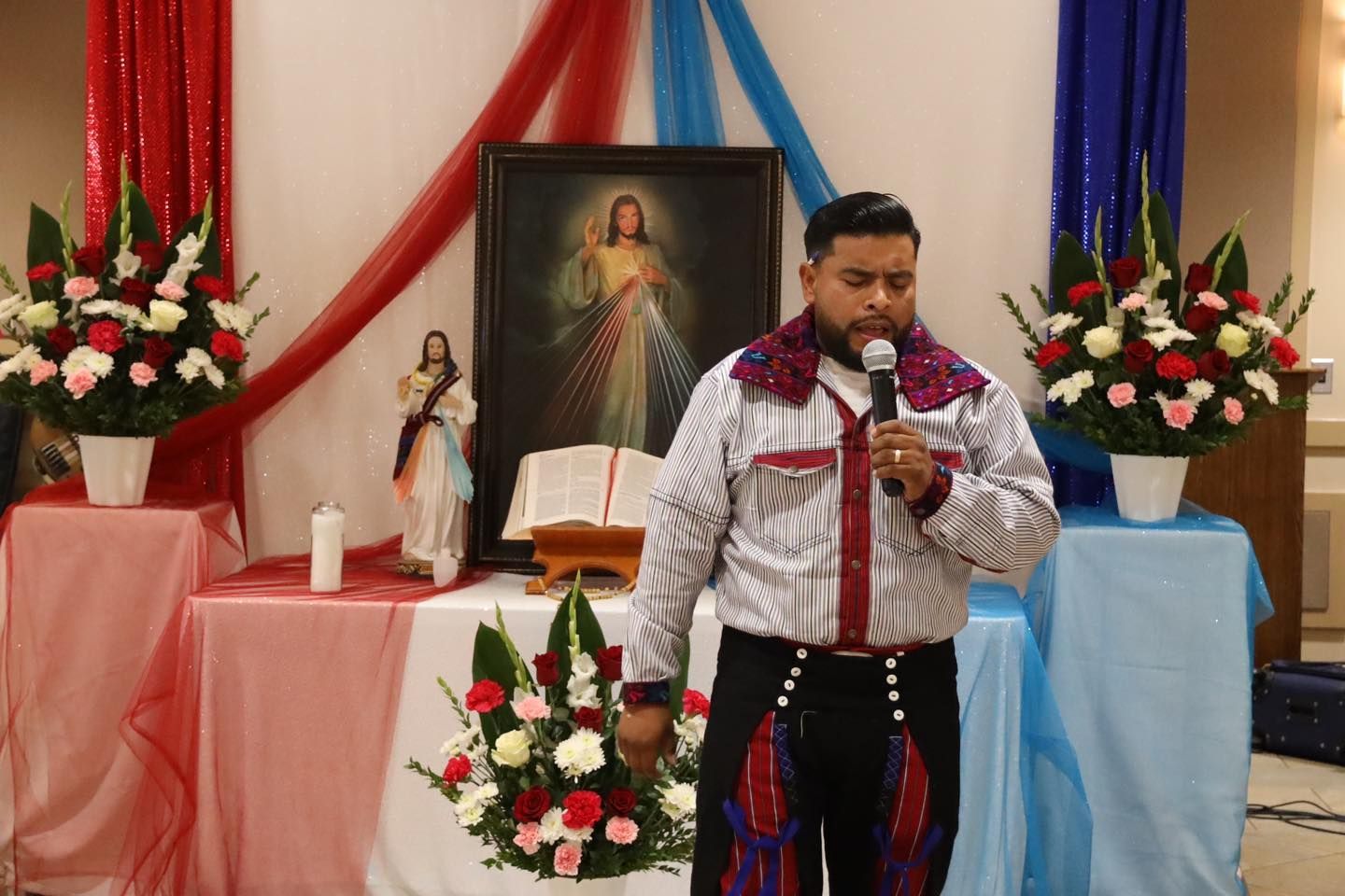 Man speaking into a microphone on a decorated stage with flowers, drapes, and a religious painting behind him