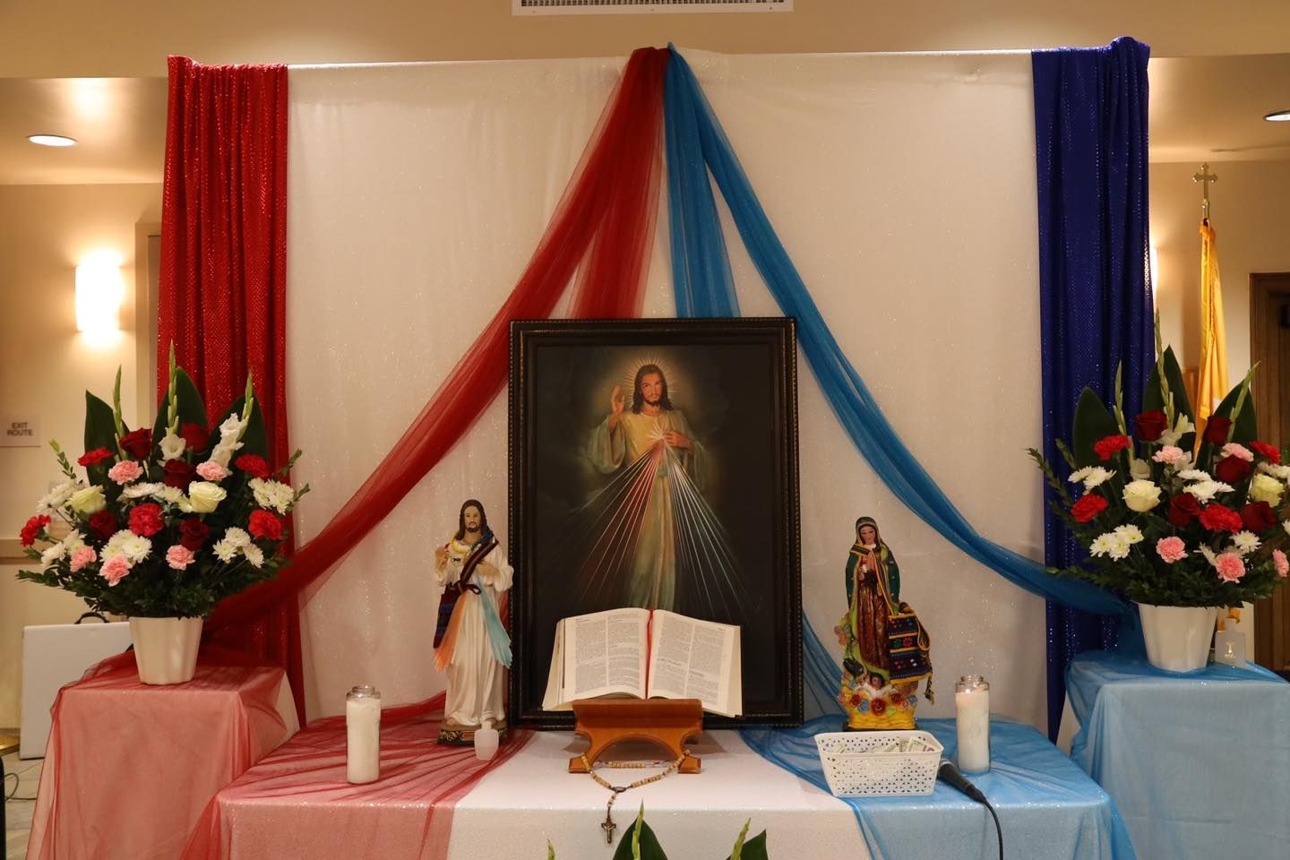Decorated altar with framed Sacred Heart image, statues, flowers, candles, and red and blue drapes