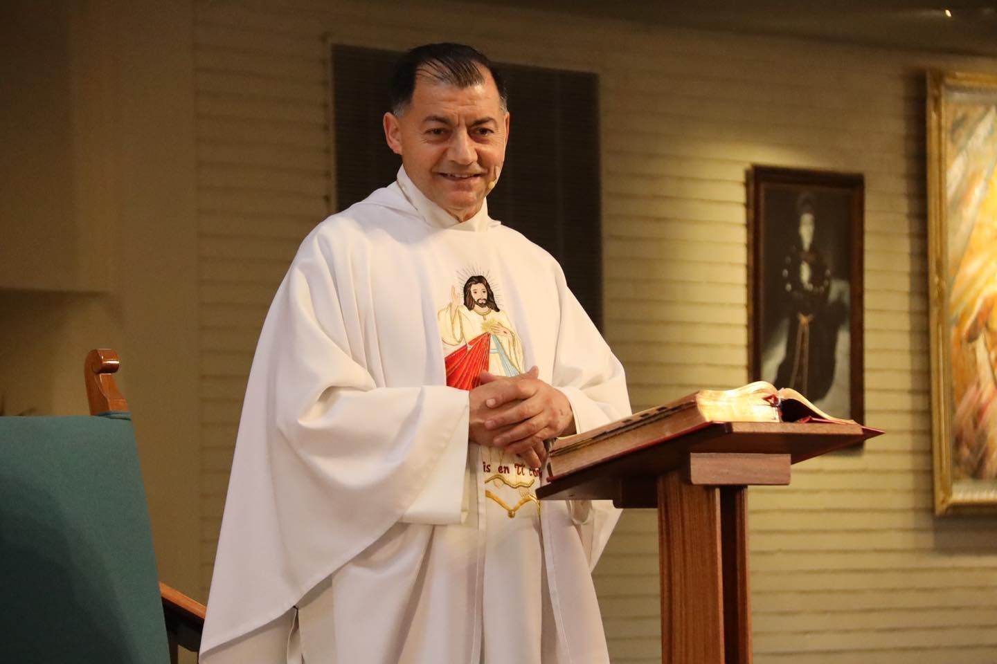 Priest in white robes stands at a lectern during a church service, smiling beside an open book.