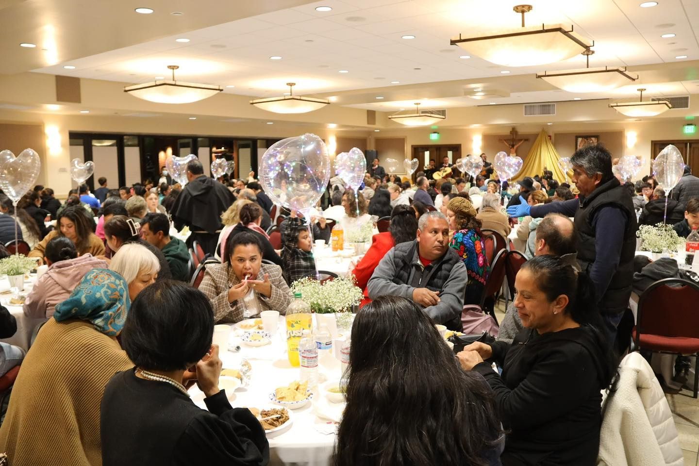 People seated at round tables in a crowded banquet hall, eating and talking under warm ceiling lights.