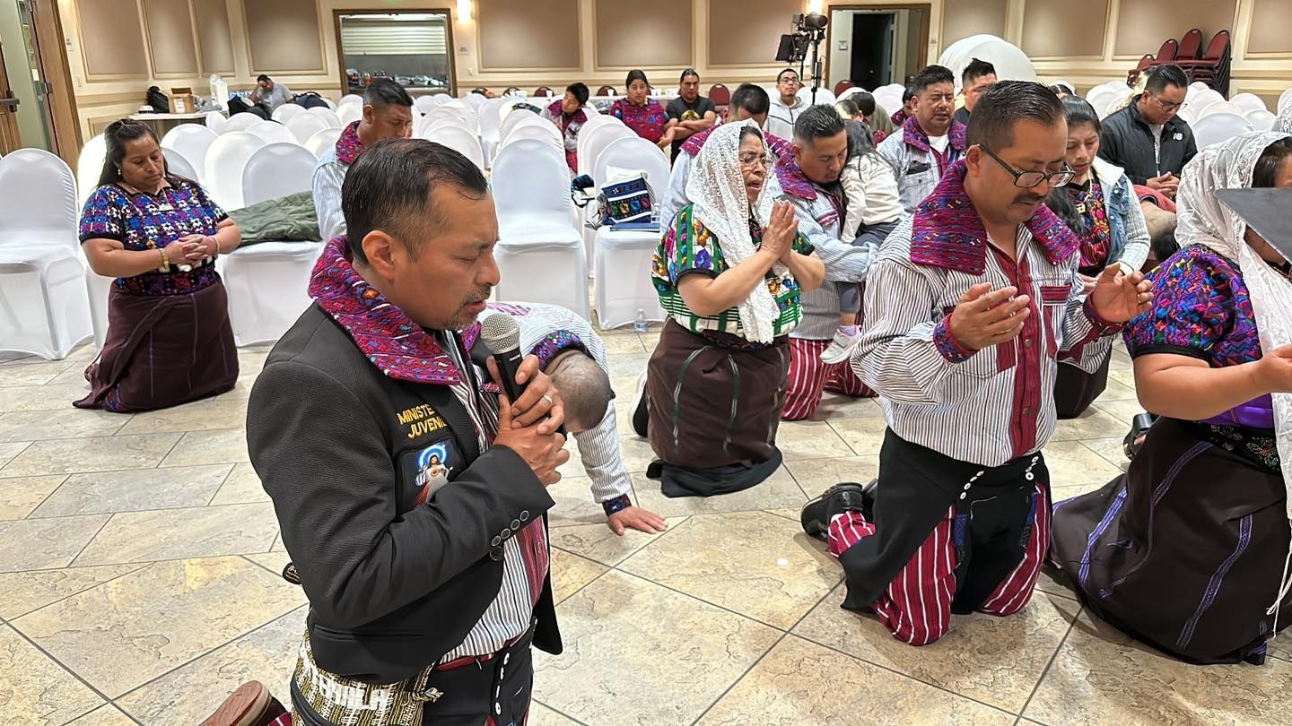 People kneeling in a ceremonial dance indoors, wearing floral leis and patterned outfits.
