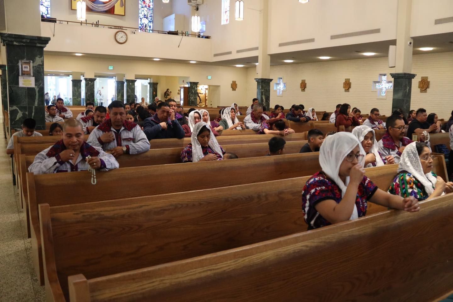 People seated in wooden pews inside a church, many wearing white head coverings.
