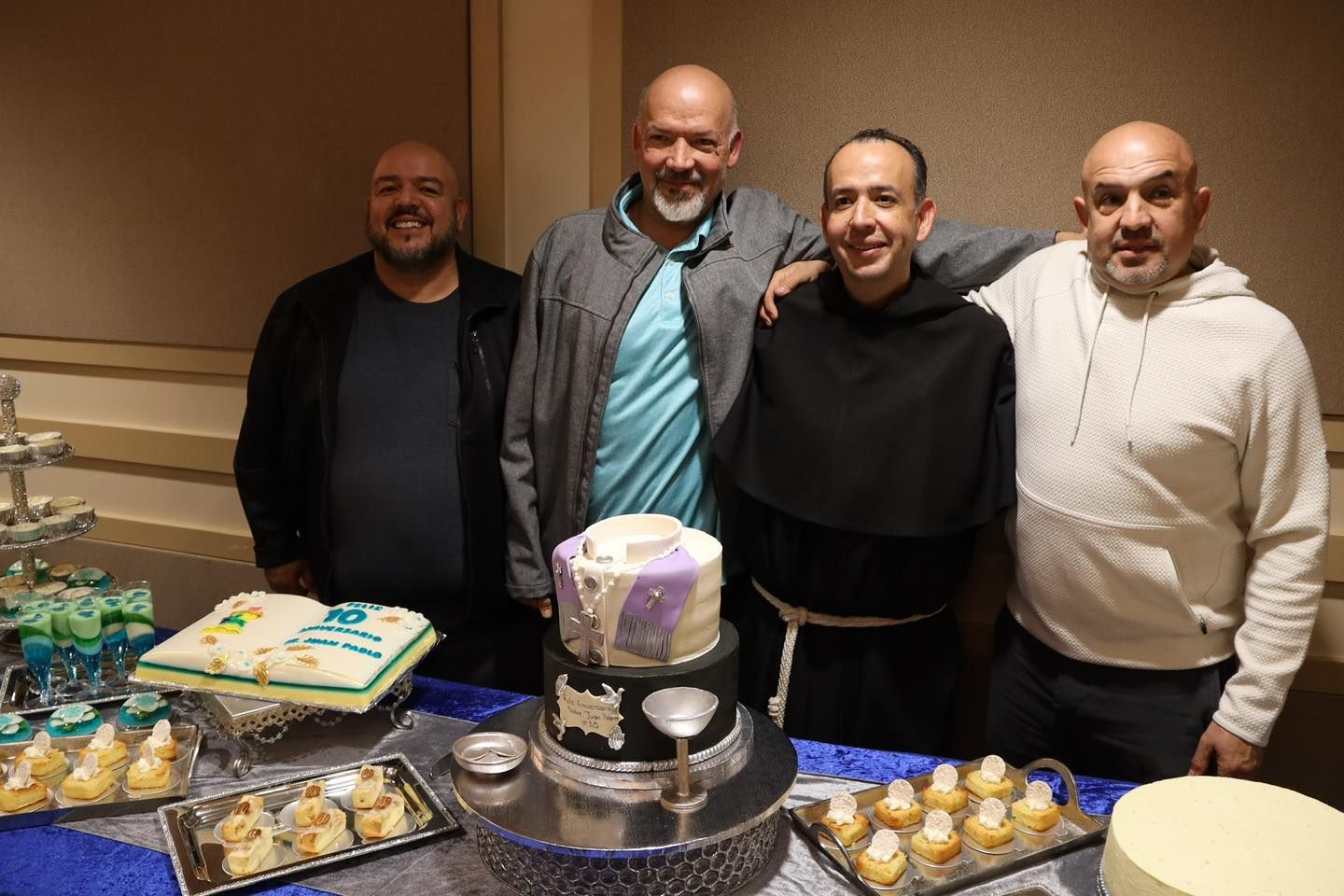Four people posing behind a birthday dessert table with cake, cupcakes, and a tiered centerpiece.
