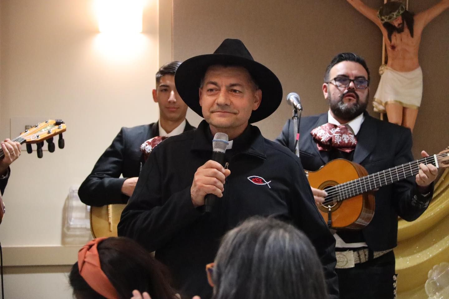 Man in a black cowboy hat speaking into a microphone while musicians play guitar behind him at an indoor event