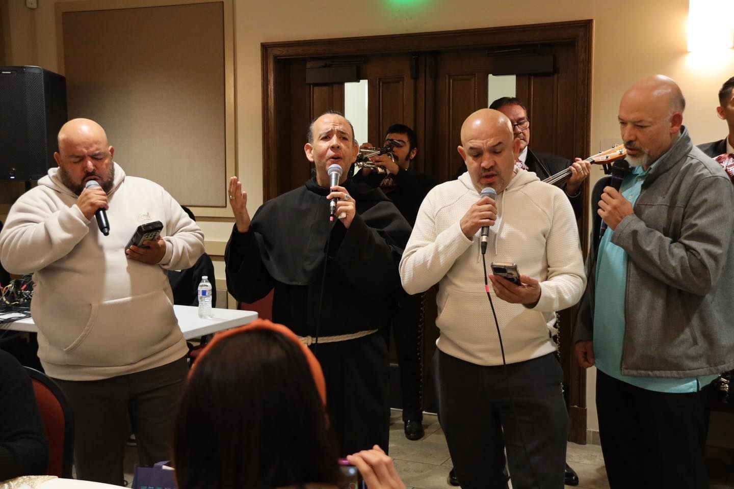 Four men singing into microphones at an indoor event, with a table and audience in the foreground.