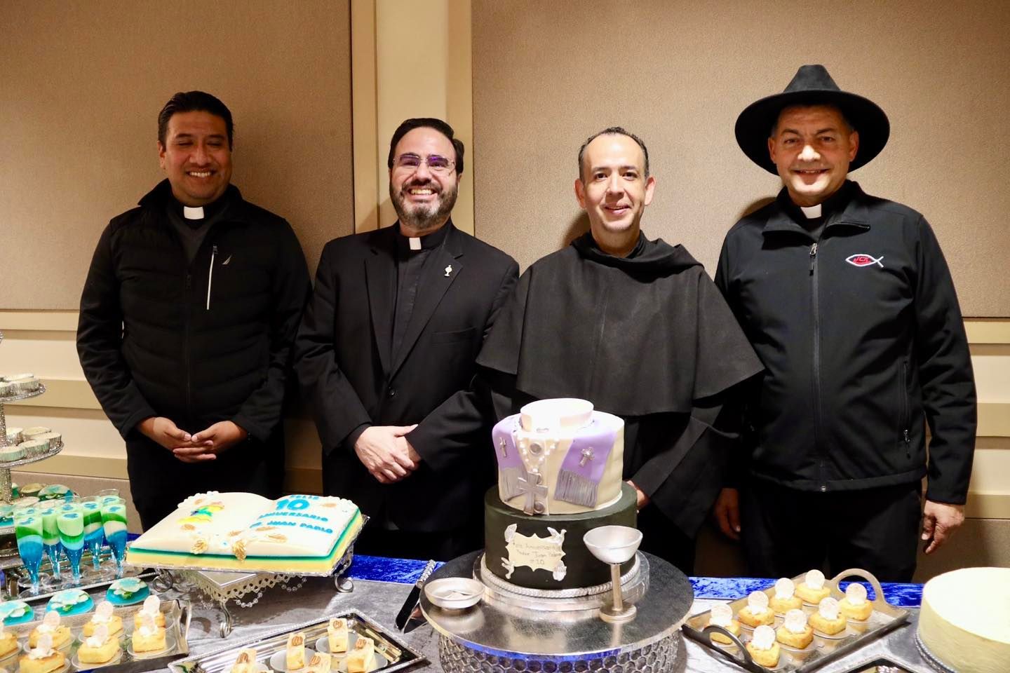 Four people stand behind a dessert table with cakes and pastries at an indoor event.