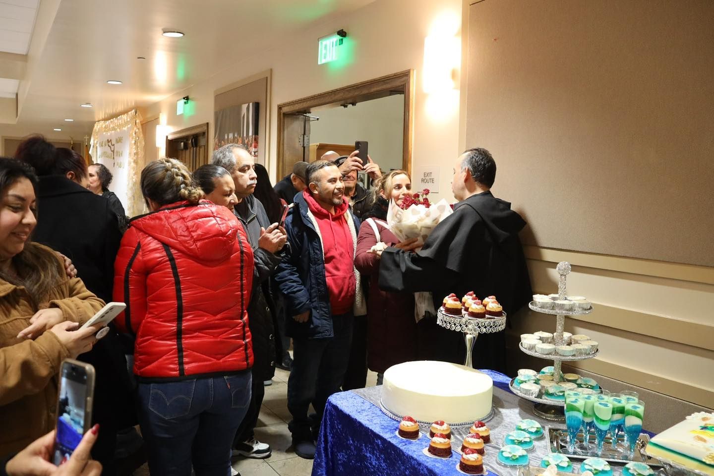People chatting at a hallway buffet table with a cake and dessert trays during a social gathering