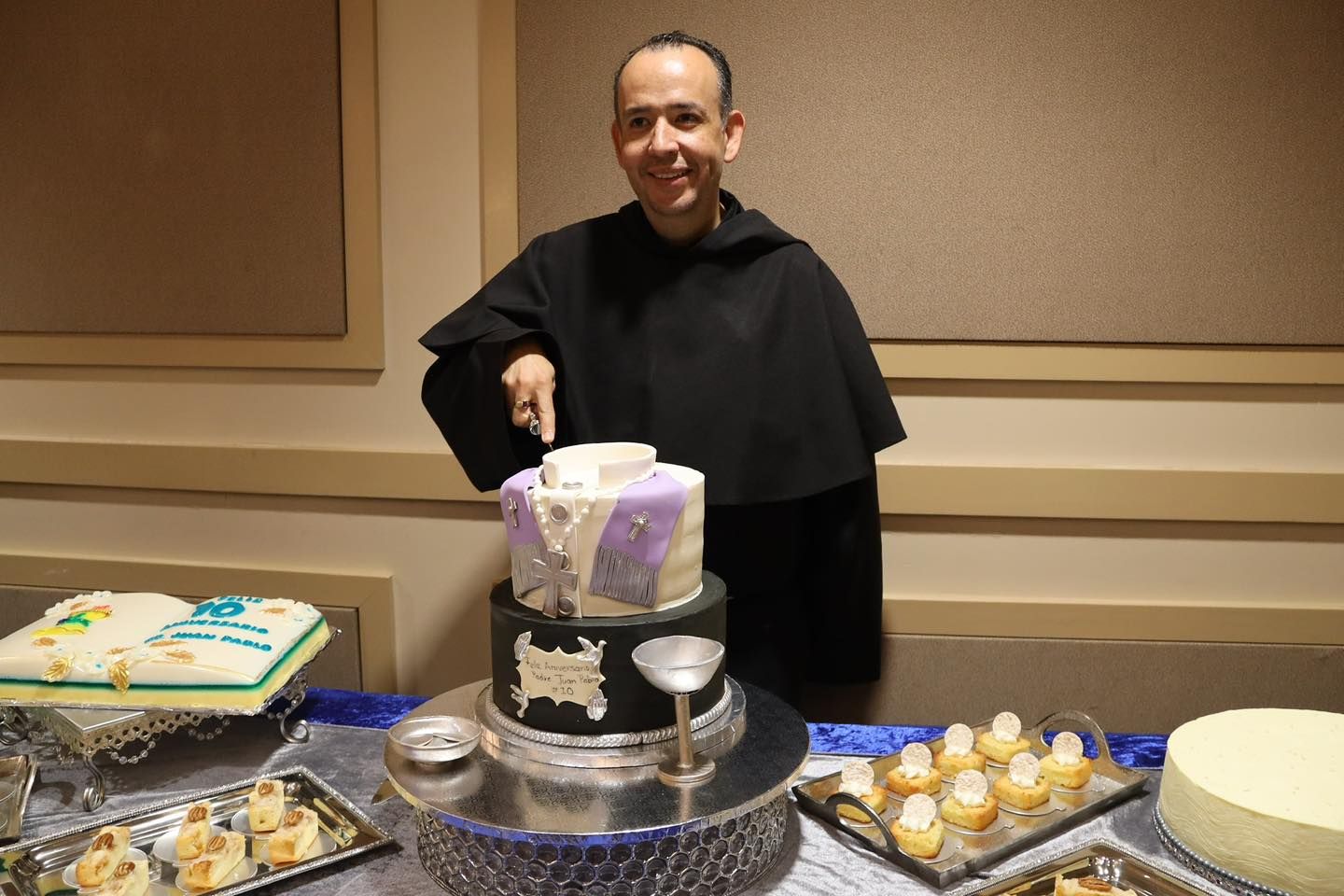 Person in a black outfit cutting a white cake at a dessert table with pastries and cupcakes