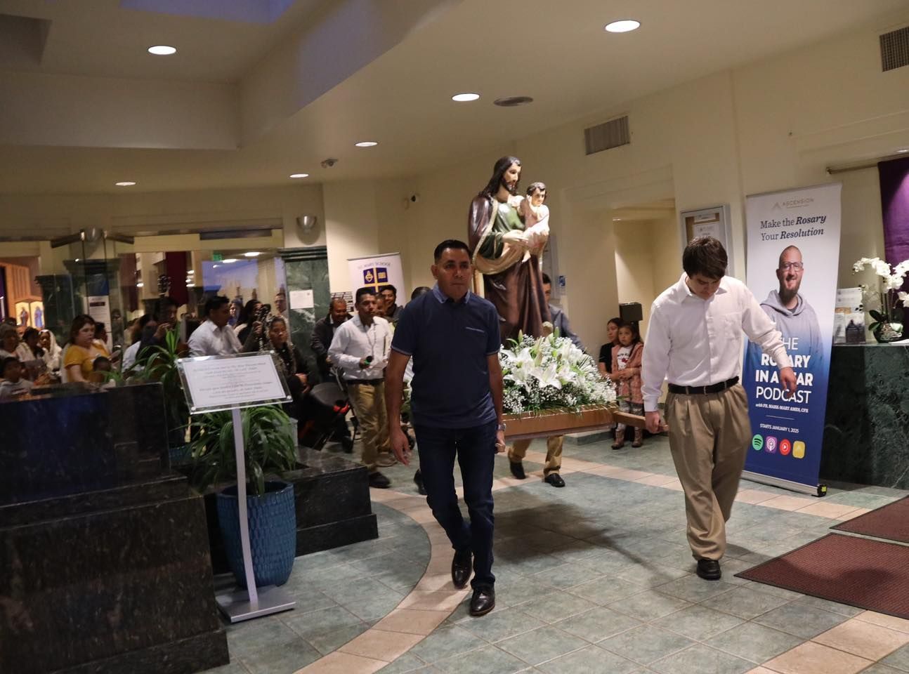 Indoor event with people carrying a decorated platform and floral arrangement through a lobby