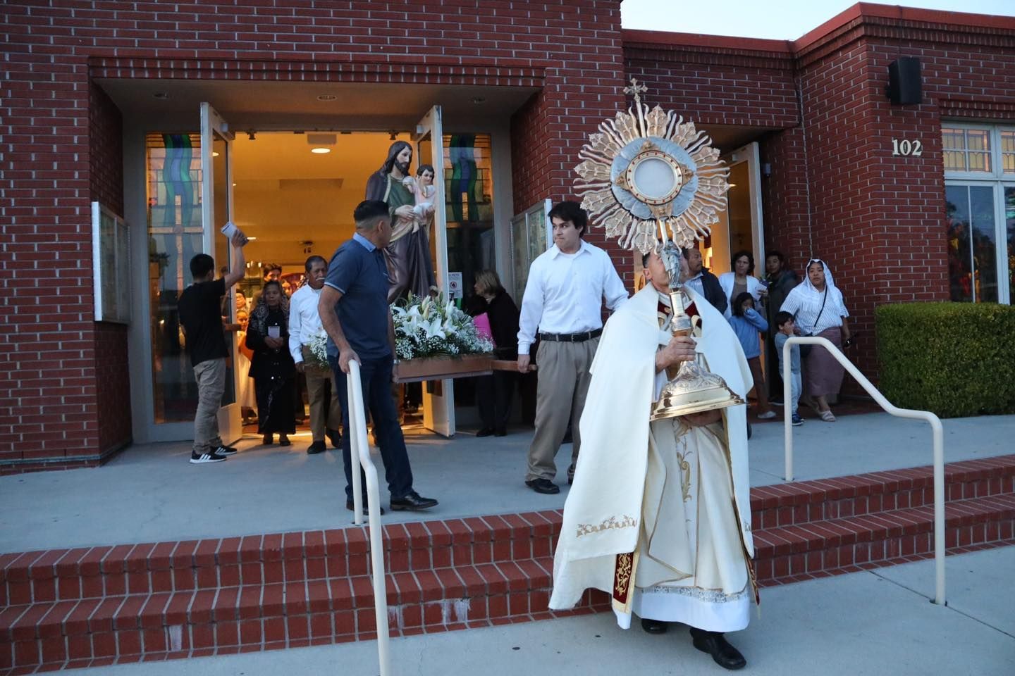 A priest in white robes holds a gold monstrance outside a brick church entrance.