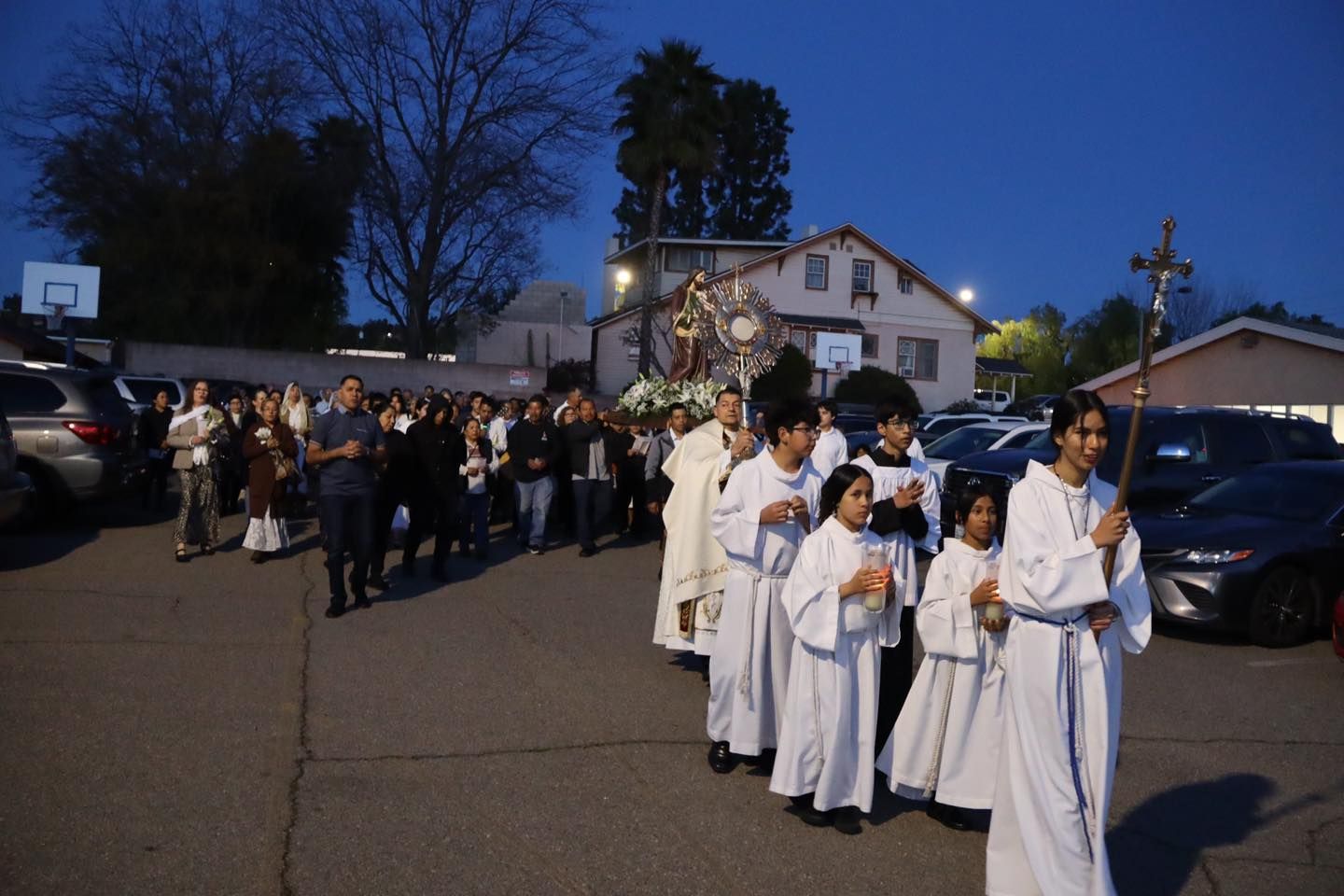 Clergy in white robes lead a candlelit procession at dusk through a parking lot near a church.