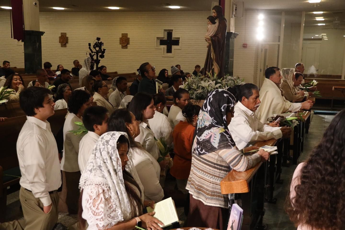 People standing and praying inside a church, with candles and a cross on the wall.