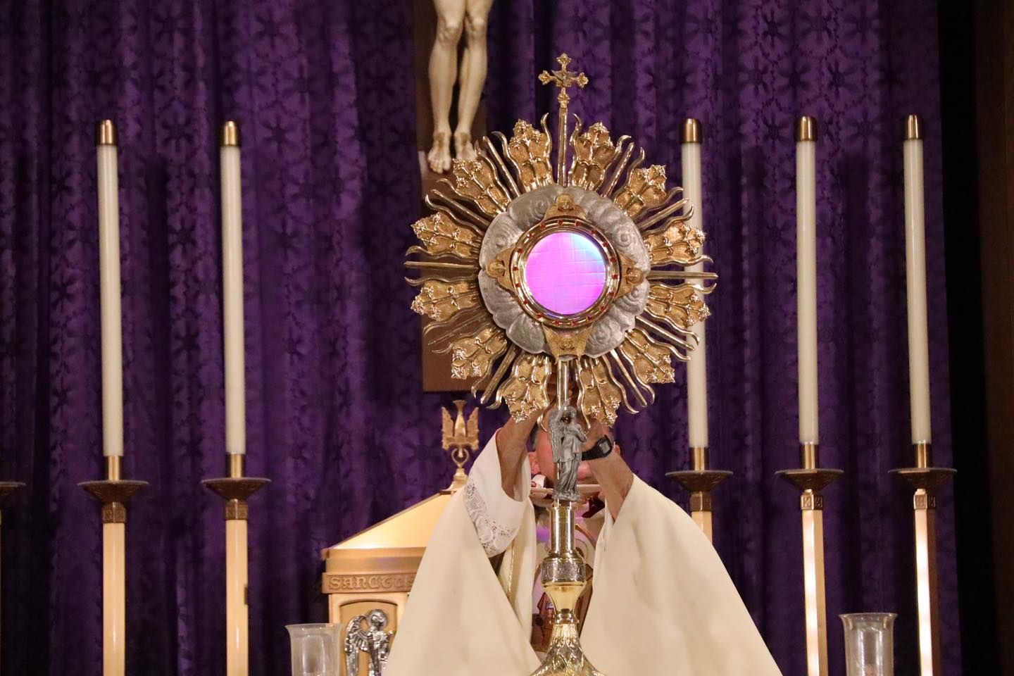 Monstrance on a church altar before a purple drape, framed by tall candles and a crucifix above