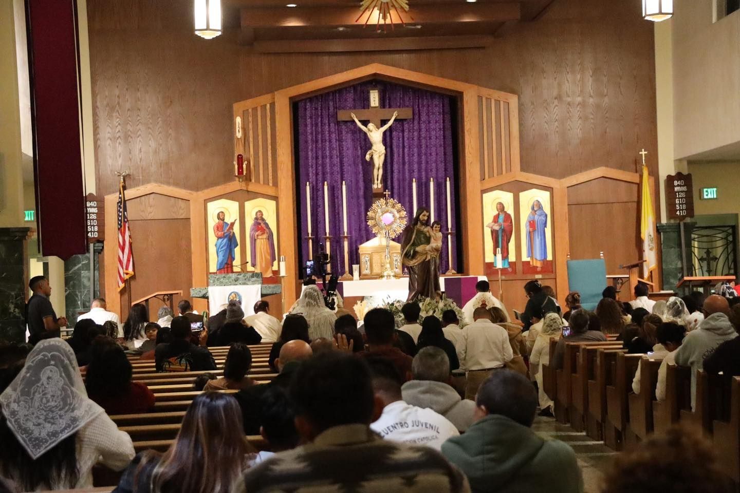 Congregation seated in a church during Mass, facing altar with crucifix and purple drapery