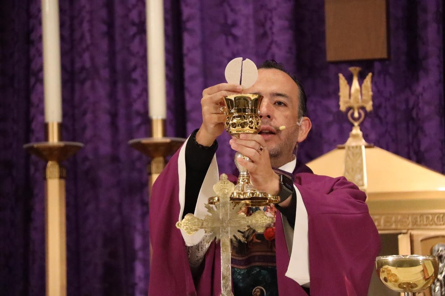Priest in purple vestments elevates a chalice during Mass before a purple curtain backdrop