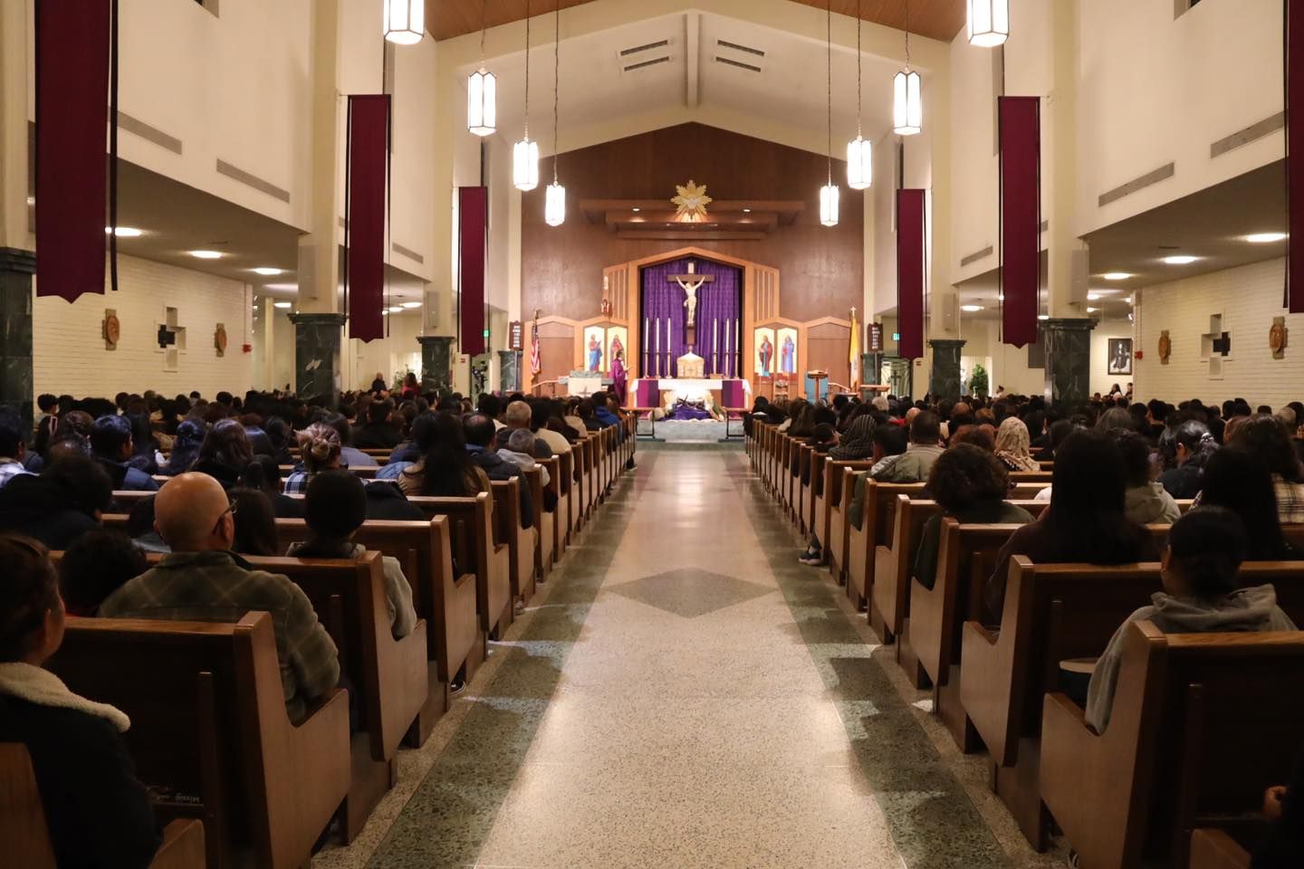Church interior with people seated in pews, facing a lit altar with purple drapery.