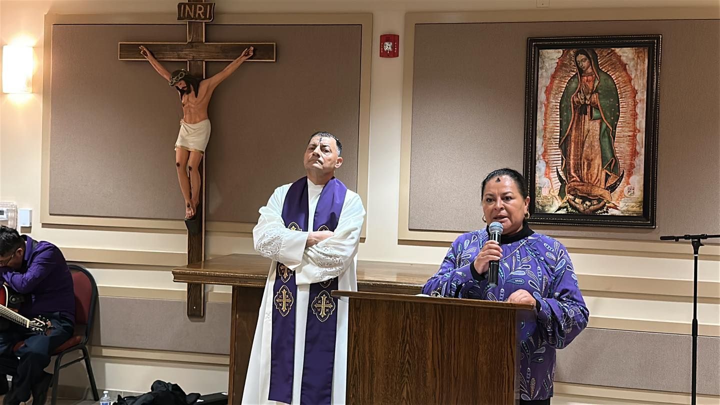 Church service with two people at the front: one at a lectern, another speaking into a microphone beneath a crucifix.