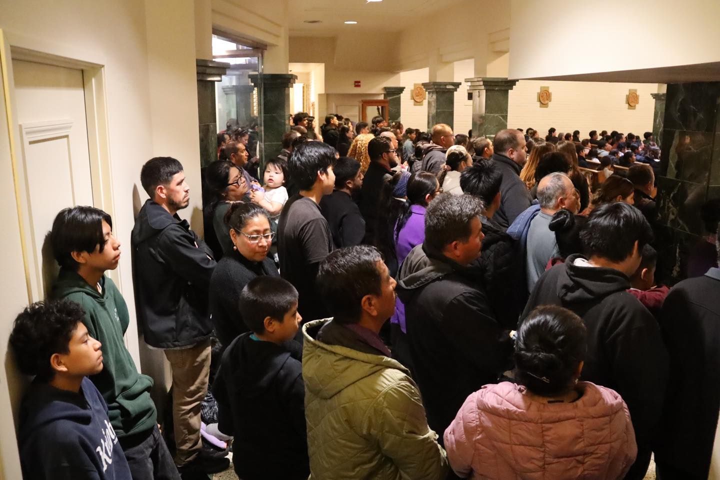 Crowded indoor hallway with people gathered closely together, many facing a doorway or speaker.