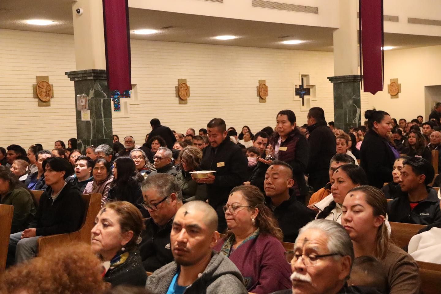 Crowded indoor gathering with seated and standing attendees in a hall, many facing forward attentively.