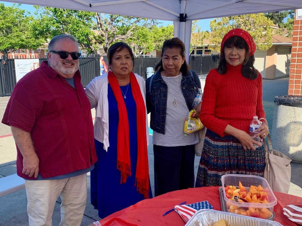 Four people stand under a canopy beside a table with drinks and snacks, posing outdoors.