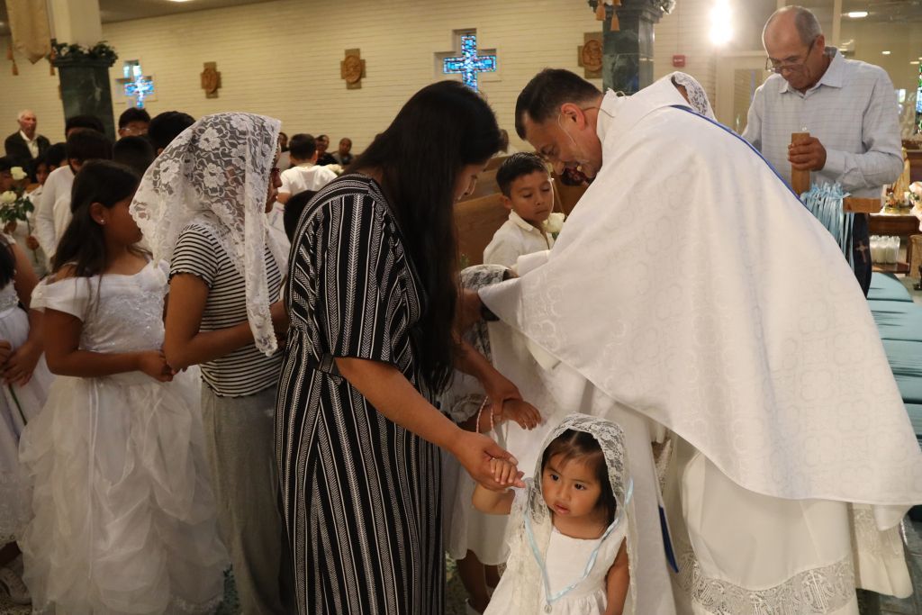 Priest in white robe blessing a child during a church ceremony with families gathered nearby