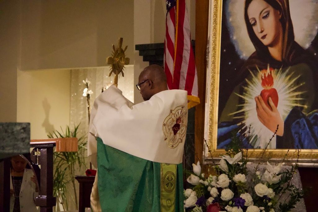 Priest in green vestments with raised white cloth at church altar beside Marian portrait and U.S. flag