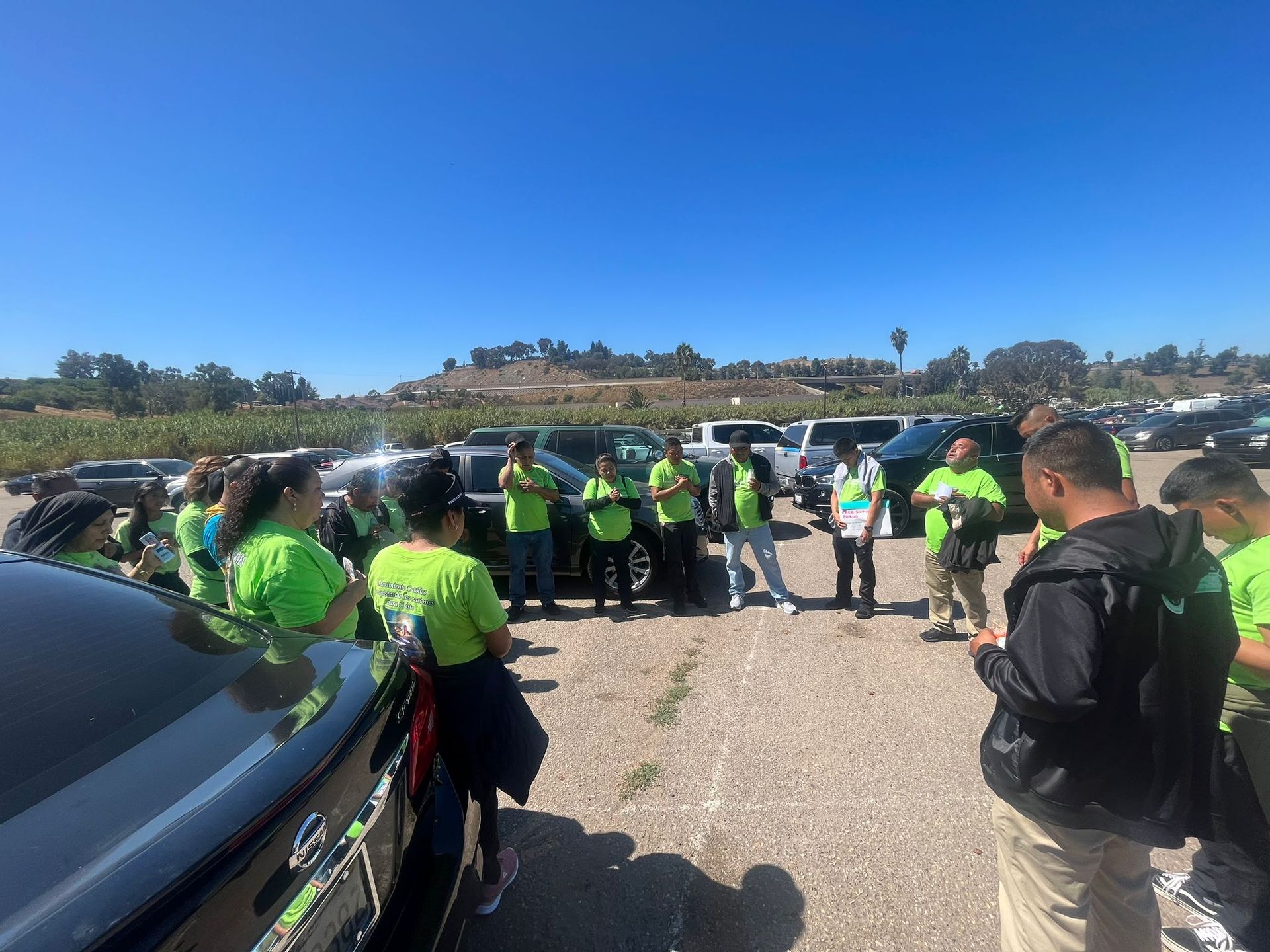 Group of people in neon safety vests gathered beside parked cars on a gravel lot under a clear blue sky
