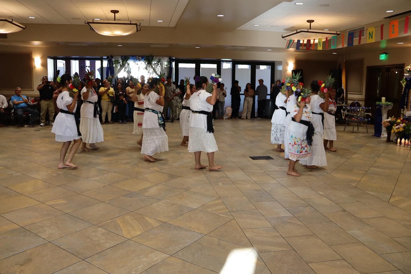 People in white outfits dancing in a bright hall with a seated audience in the background