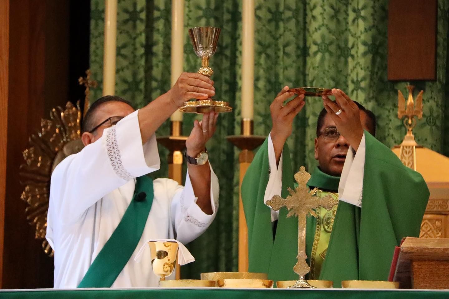 Two clergy in green vestments raise chalices at an altar during a church service