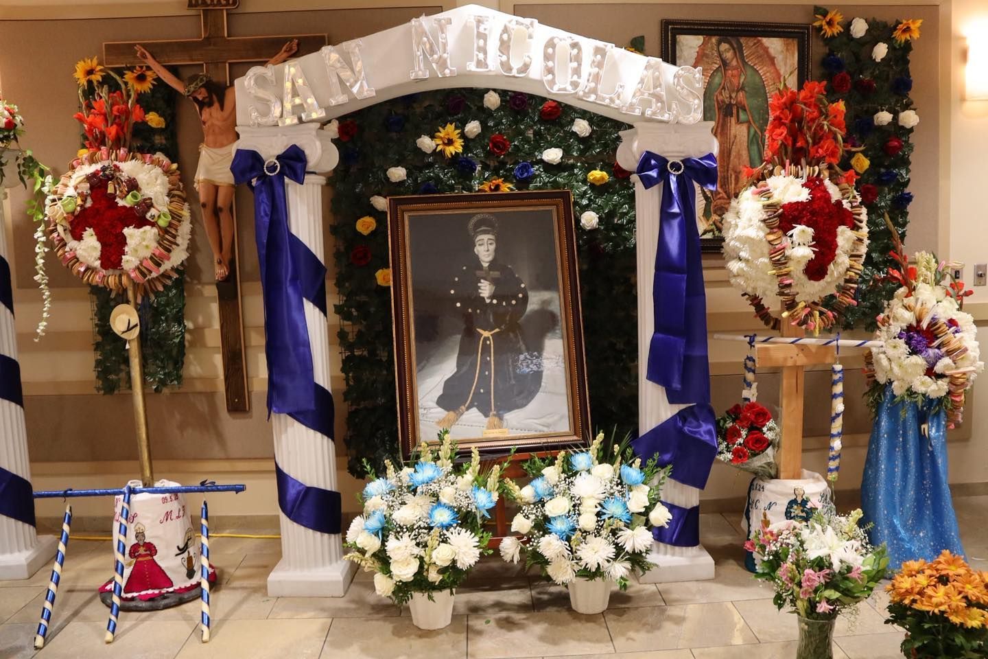 Memorial altar with framed portrait, blue and white drapes, floral wreaths, and candles at an indoor ceremony