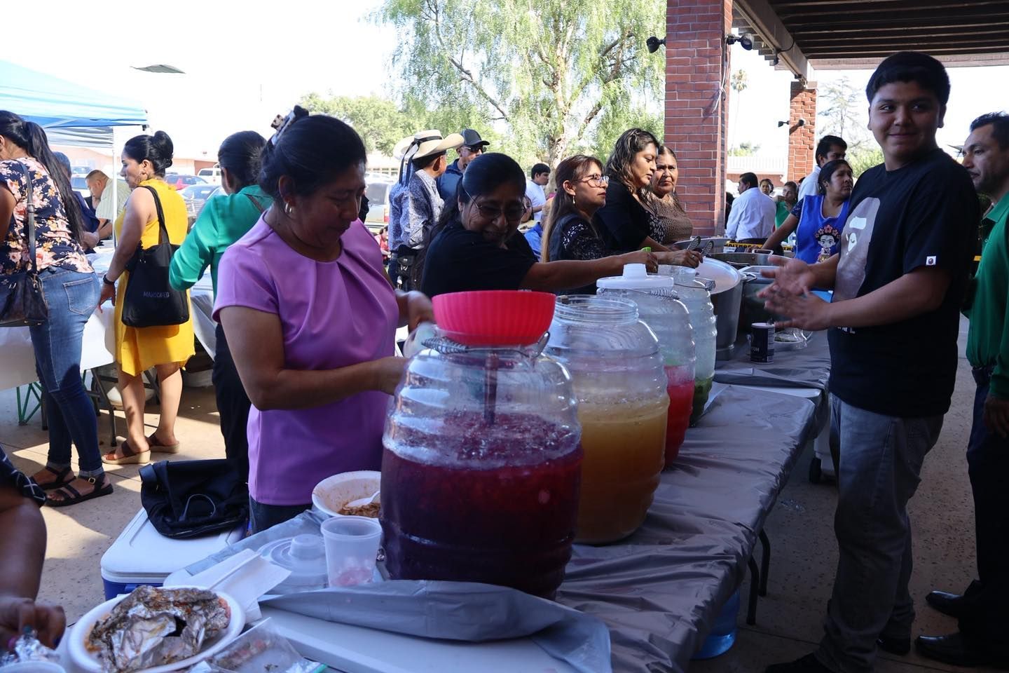 People serving drinks from large jars at a crowded outdoor food stand.