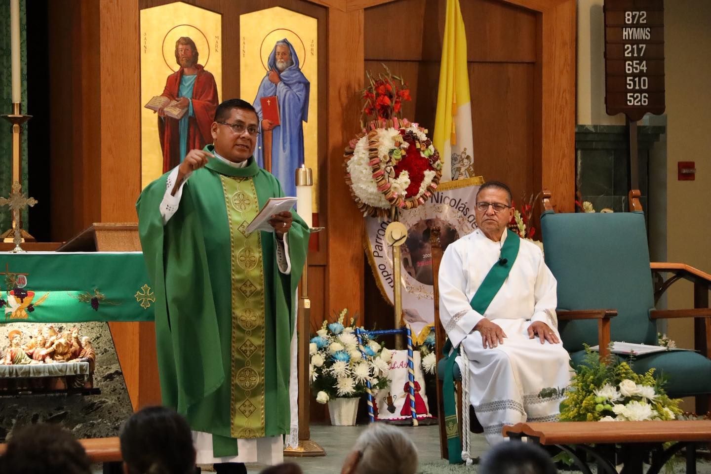 Priest in green vestments speaks at altar beside seated clergy in white, inside a church sanctuary