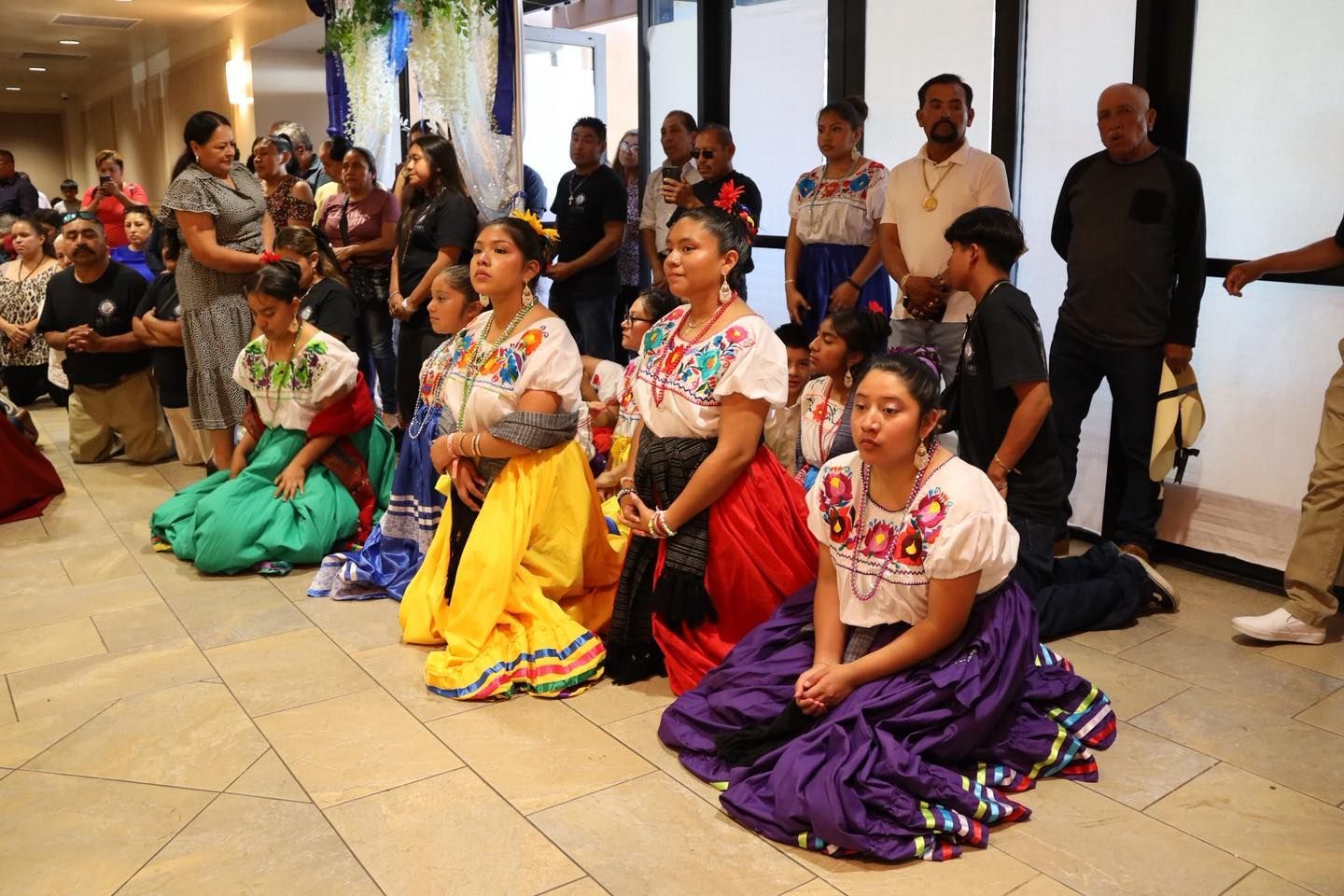 Four dancers in colorful traditional dress kneel in a row during a performance with an audience behind them.
