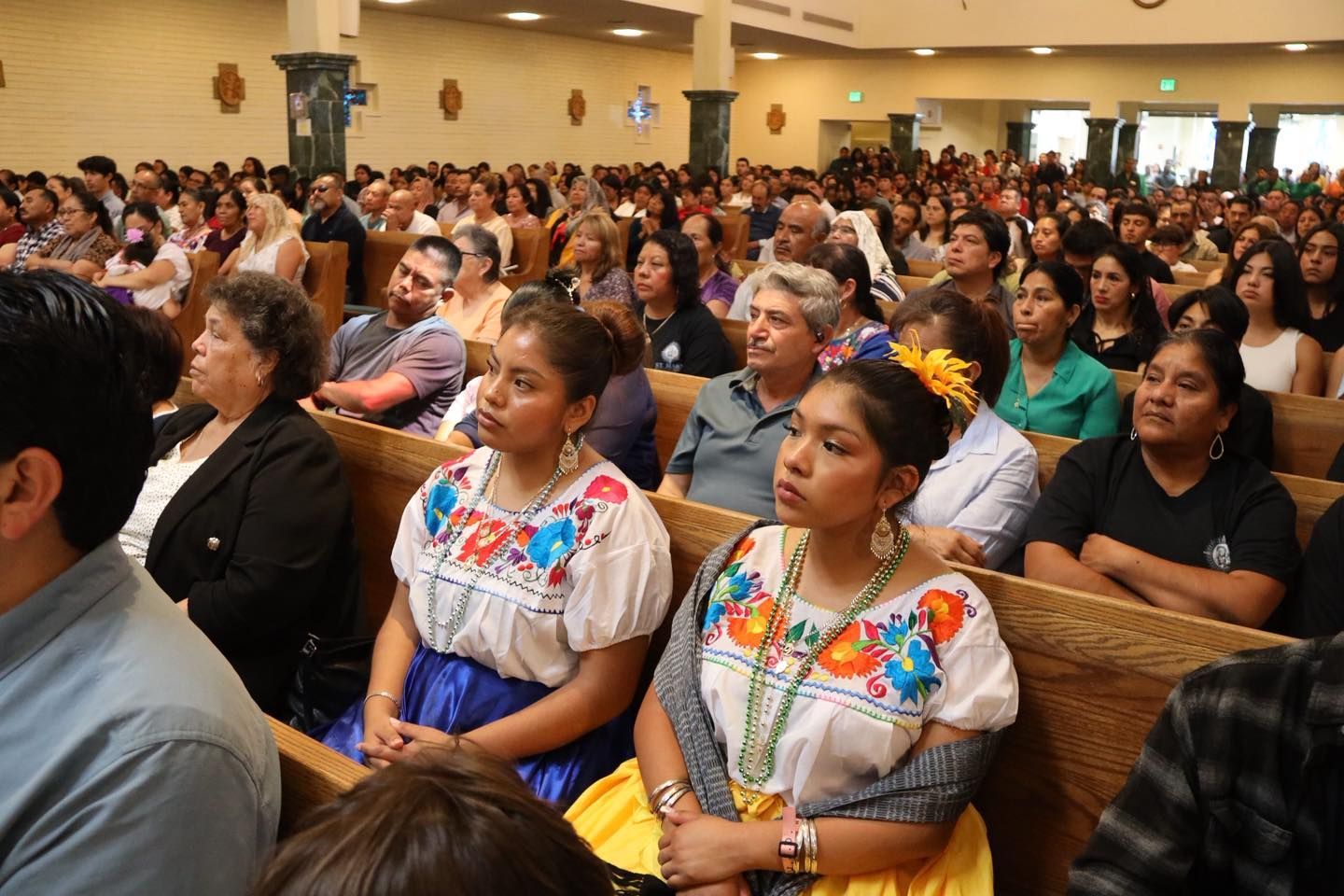 Audience seated in a church hall, with several women in colorful embroidered blouses in the foreground.