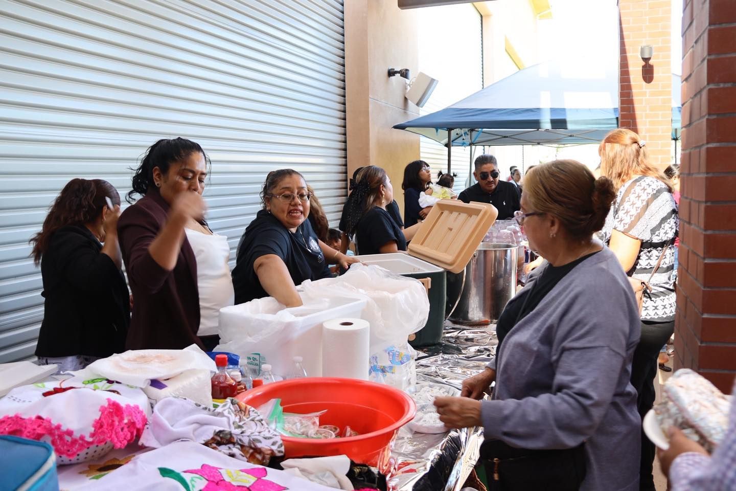 People at an outdoor table with white bags and supplies, chatting near a closed storefront.