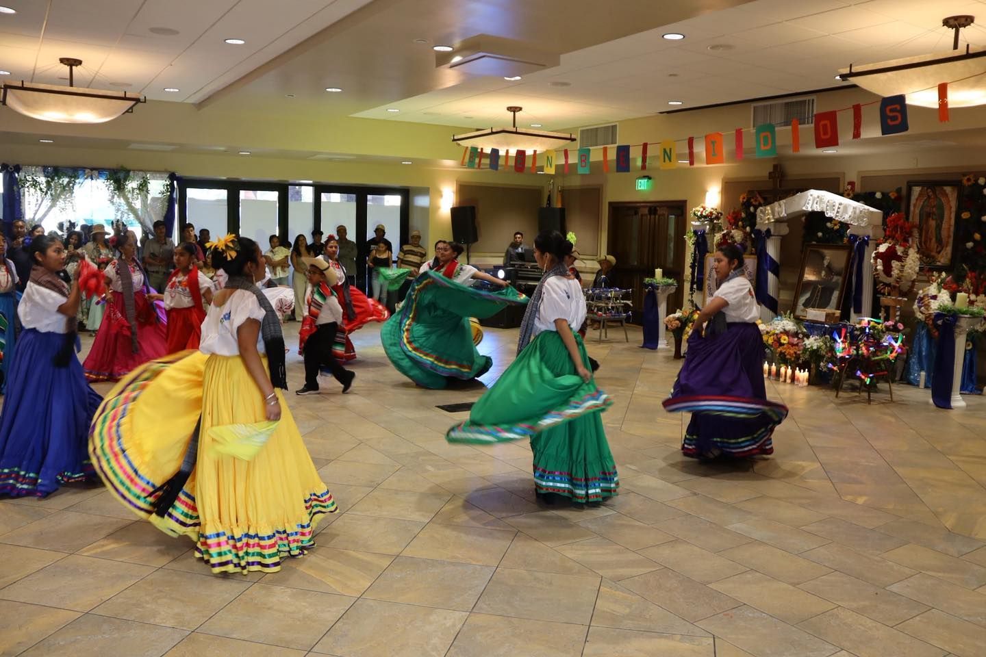 Dancers in colorful traditional skirts perform in a banquet hall, with spectators seated around them.