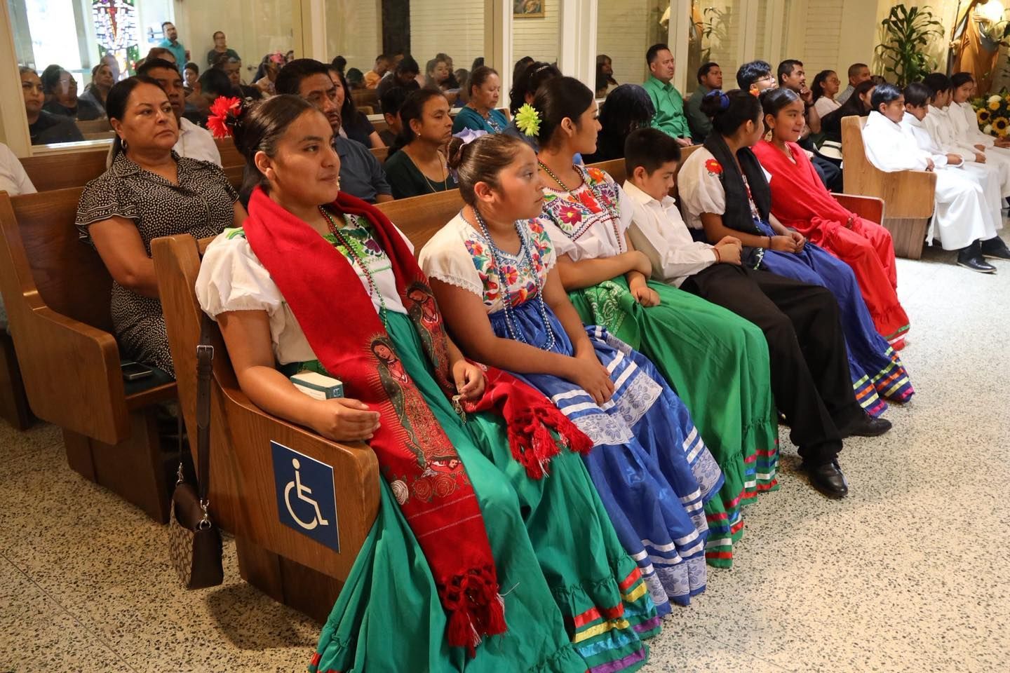 People seated in a church pew, wearing colorful traditional dress and red sashes, during a ceremony
