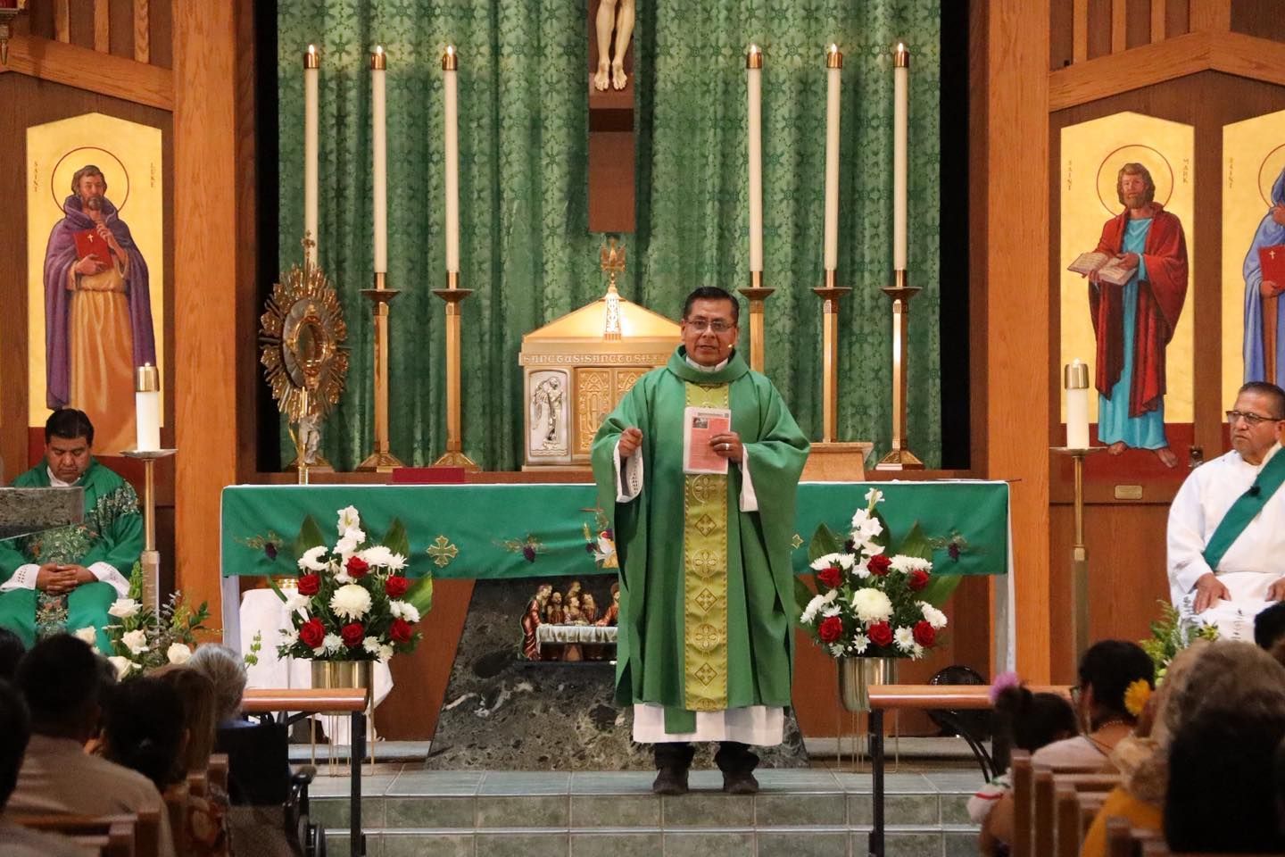 Priest in green vestments speaking at the altar during church Mass, with candles and floral arrangements behind him