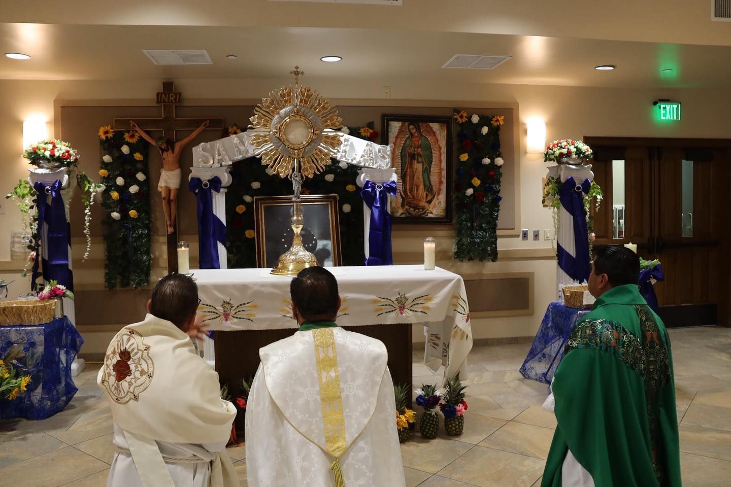 People kneel before a decorated altar in a church, with candles, flowers, and a gold sunburst centerpiece.