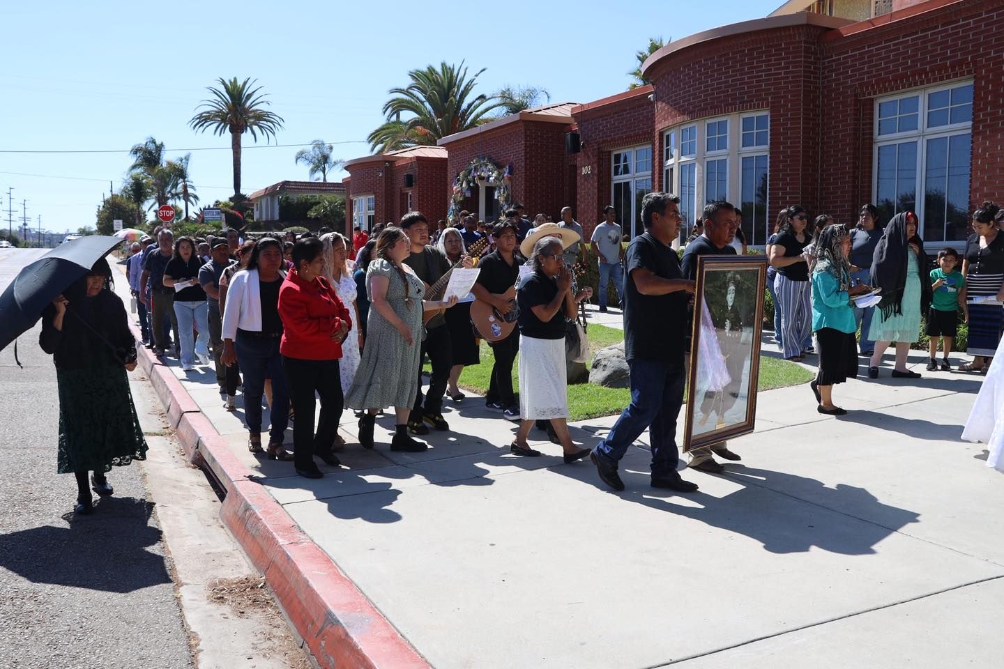 People standing in line outside a brick building on a sunny day