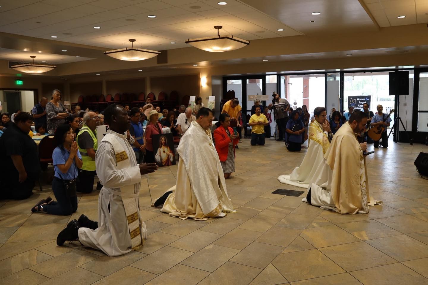 People kneeling on a tiled floor in a hall during a ceremony, with onlookers gathered around.
