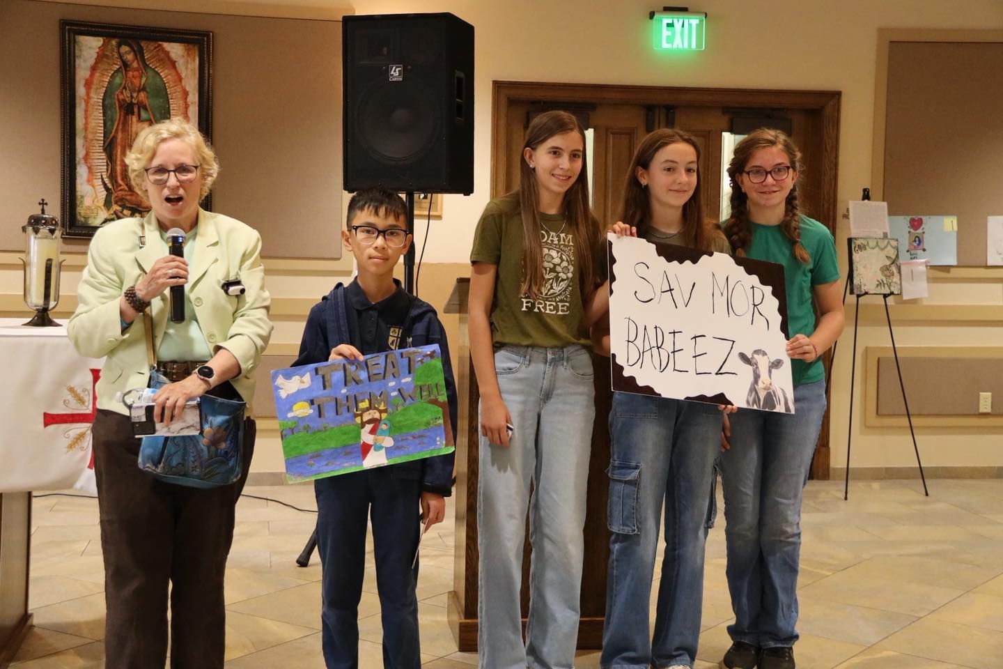 Four people stand indoors at a microphone, holding handmade signs during a presentation.