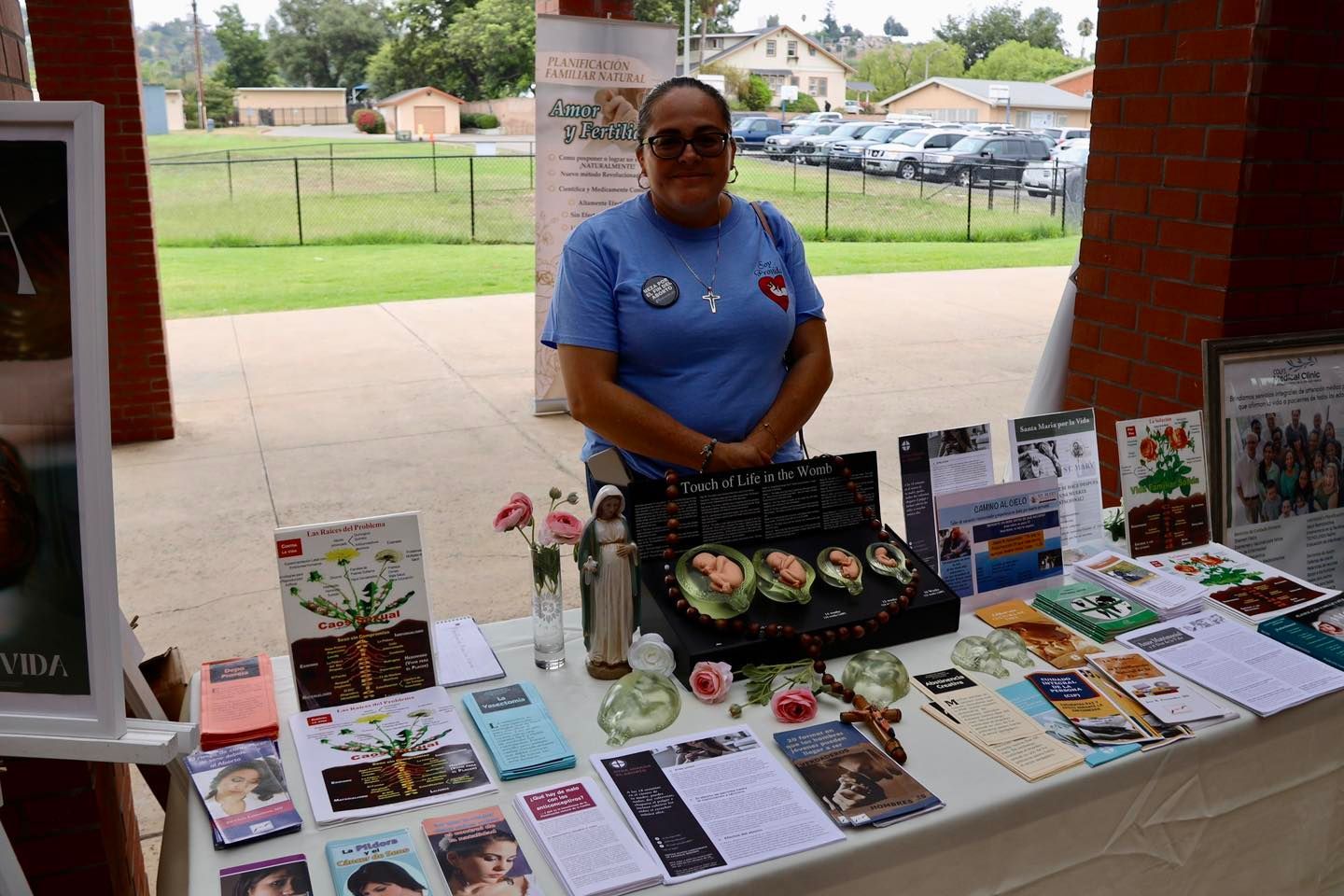Person standing behind a table of handmade crafts and books at an outdoor market stall.