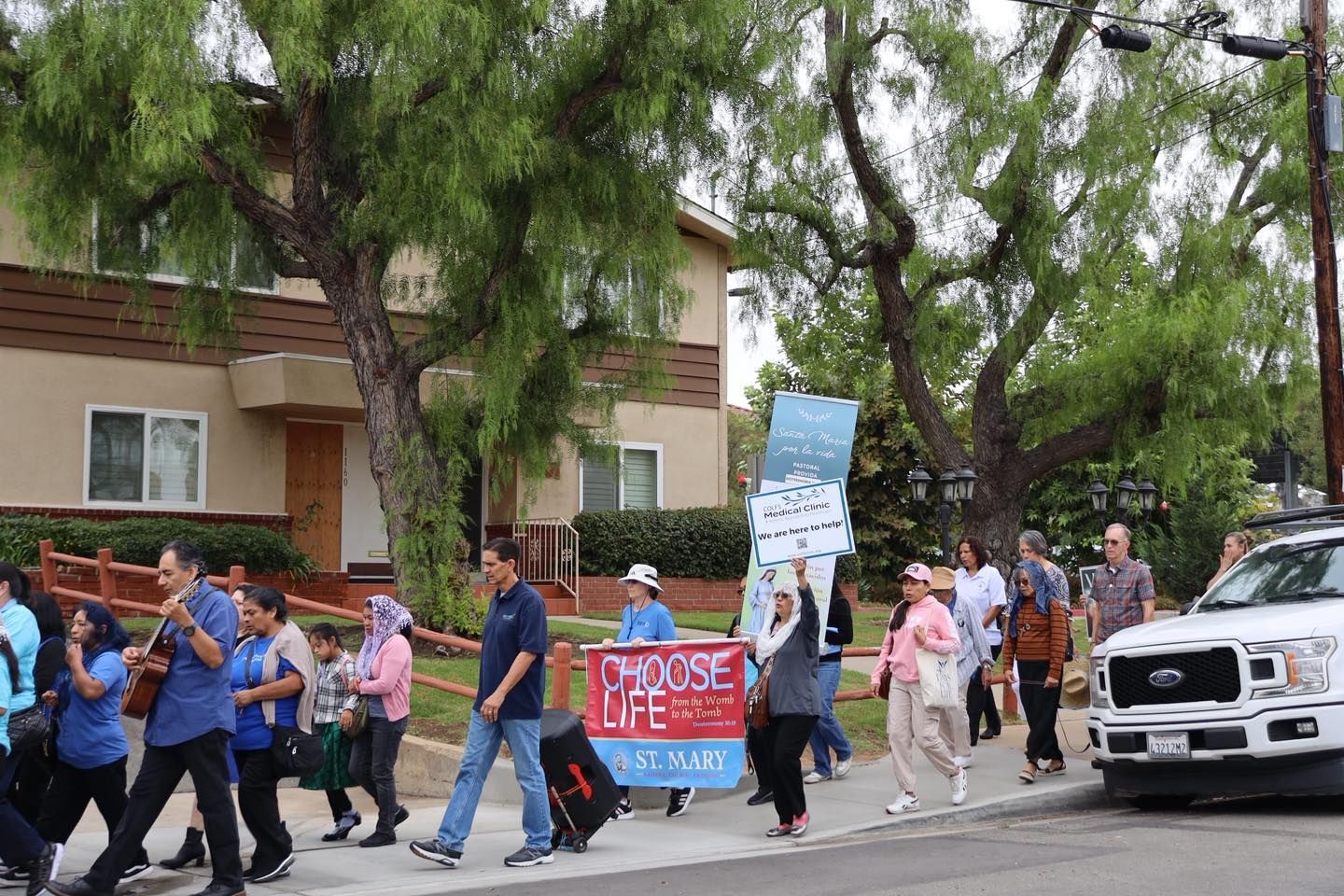 Protesters marching on a street with signs, including one reading “Protect Life,” near parked cars and trees.