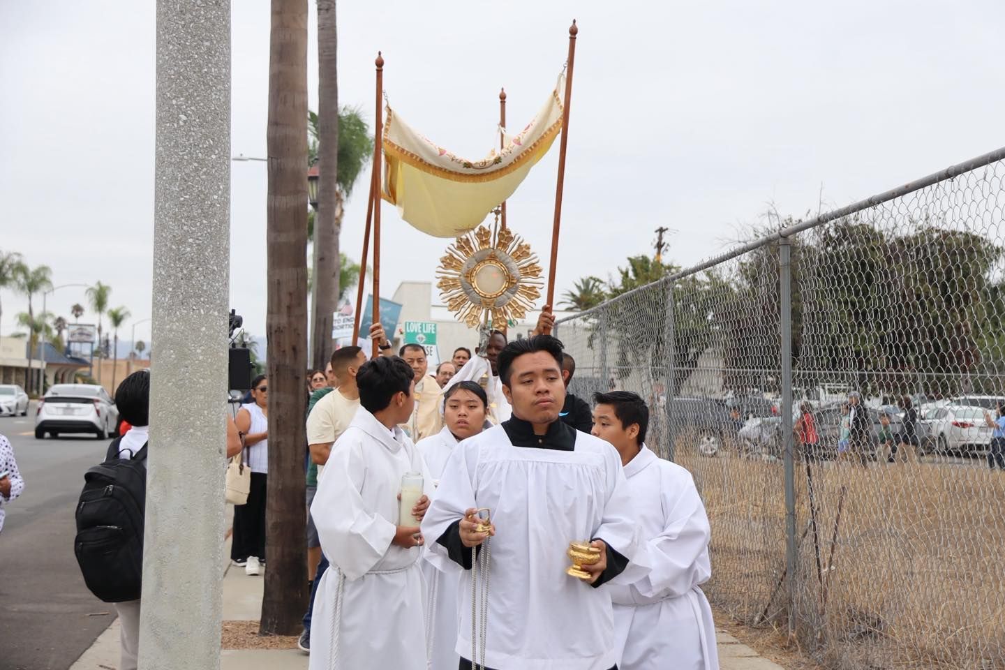 People in white robes carrying a gold ceremonial ornament outdoors by a fence