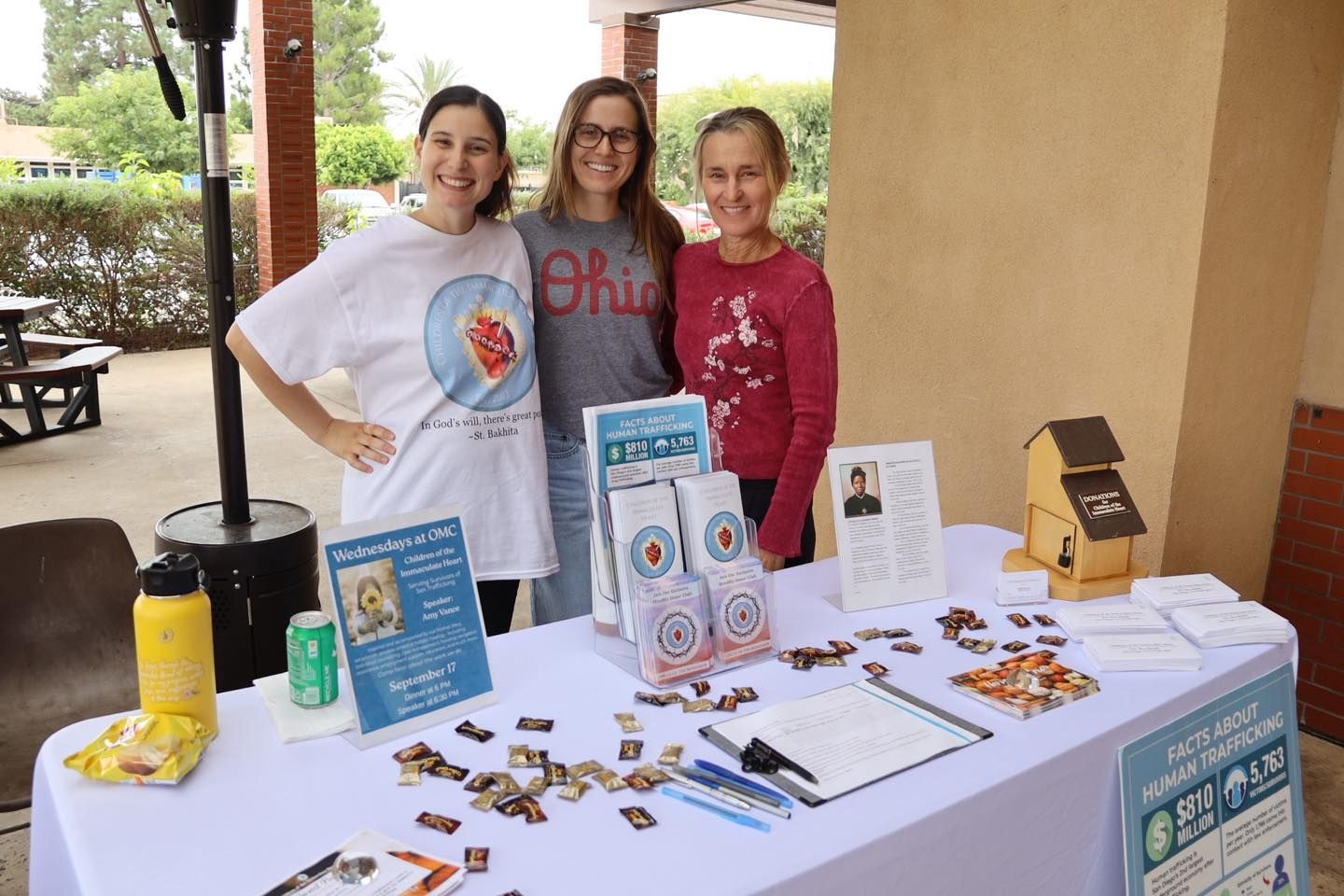 Three people at a booth display flyers and small items on a table under a covered outdoor area.