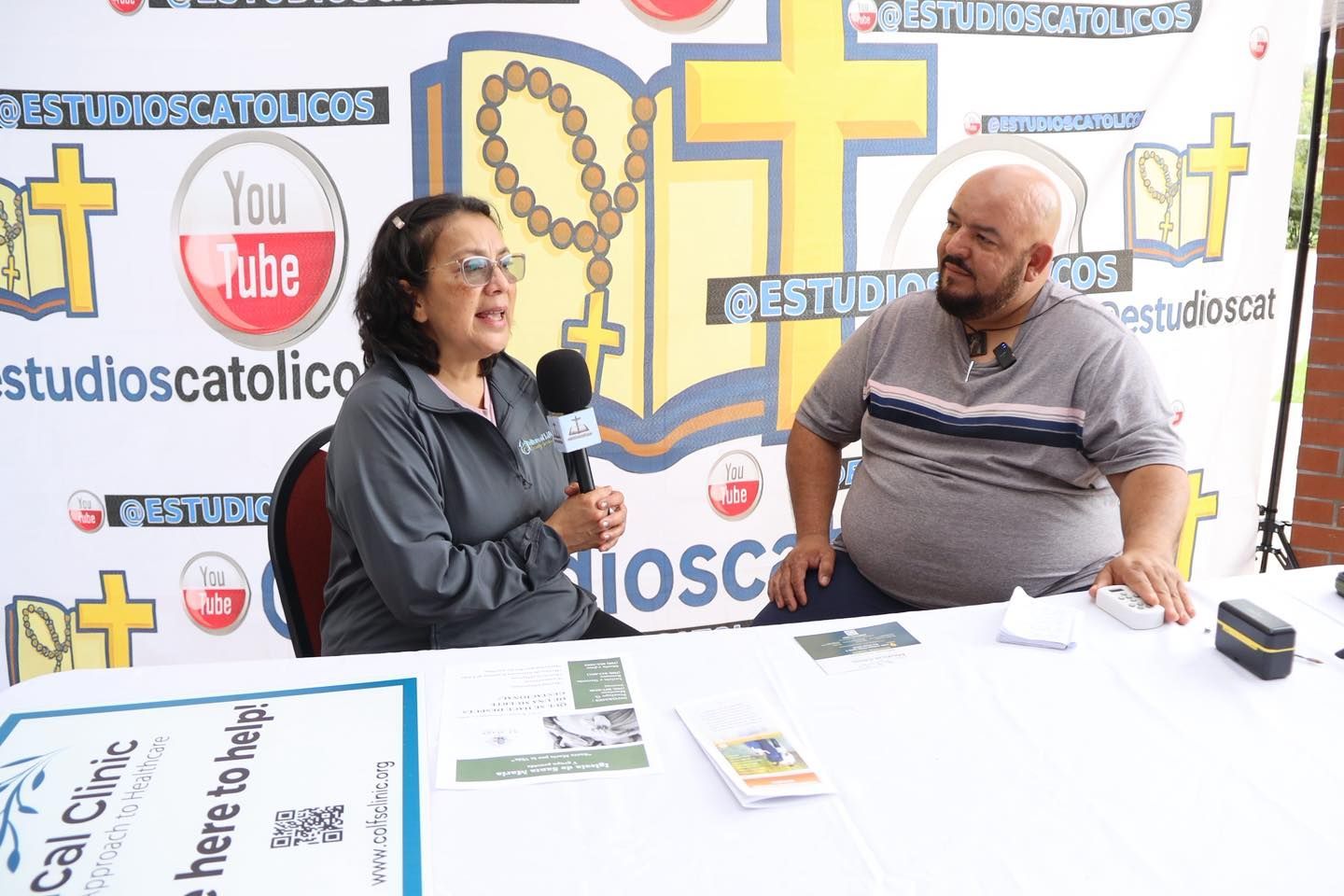 Two people talk at a table in a studio with a cross and YouTube-themed backdrop