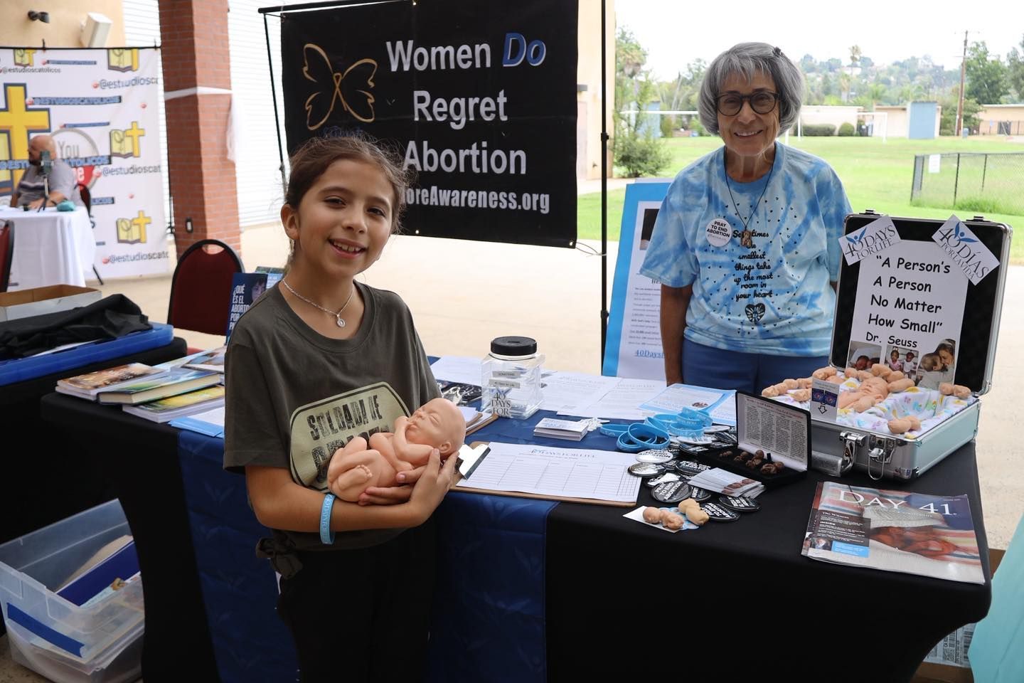Two people at a table with women’s rights and abortion protest signs and pamphlets at an outdoor event