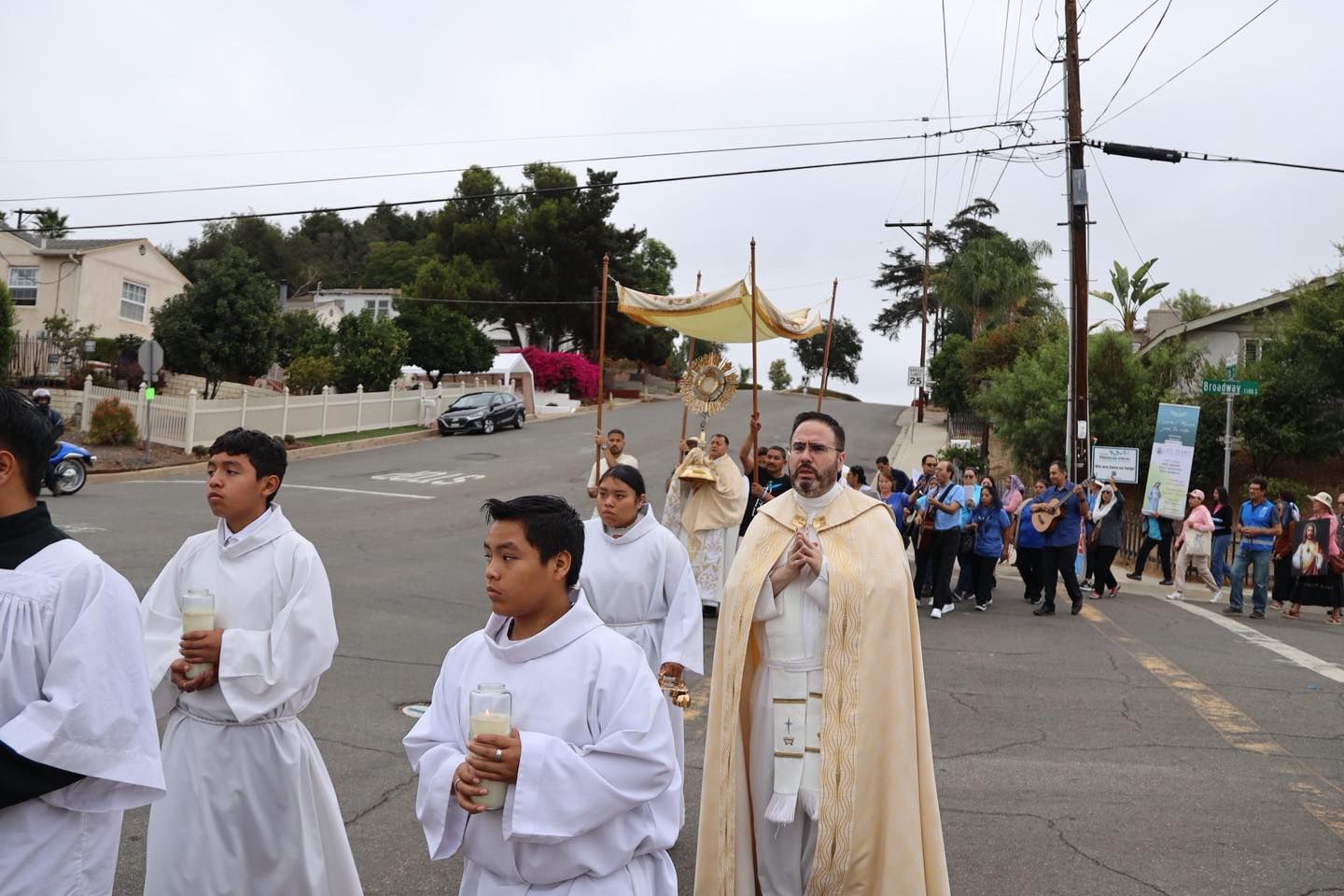 People in white robes and a priest walk in a procession along a street.