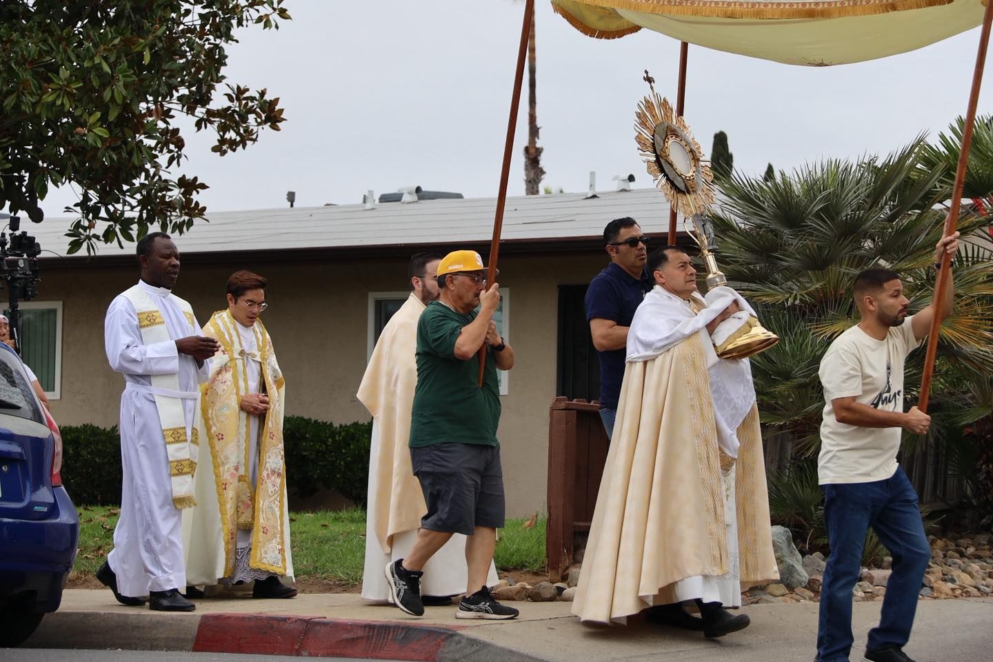 Religious procession under a canopy, with participants carrying a statue and candles along a street